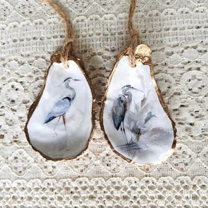 May include: Two decorative oyster shells featuring detailed illustrations of grey herons. Each shell has a gold-painted edge and a natural jute twine loop attached at the top for hanging. The interior surfaces are white with artistic bird imagery.