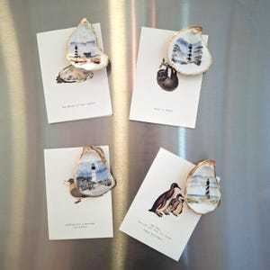May include: Four greeting cards with oyster shell magnets. Each shell is painted with a coastal scene and attached to a card with a different illustration and message. The cards are on a silver surface.
