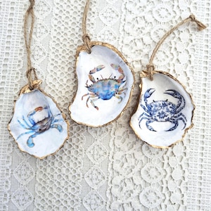 Puede incluir: Tres adornos decorativos de concha de ostra. Cada concha tiene un borde dorado y un diseño de cangrejo en acuarela en tonos azules. Las conchas están suspendidas por hilo, creando un tema costero o náutico.