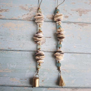 May include: Two decorative wind chimes. Each chime features a strand of stacked, cream-colored seashells, interspersed with light blue beads and gold-colored beads. A small, gold-colored bell hangs at the bottom of one chime, and a gold-colored cone hangs at the bottom of the other.