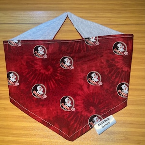 May include: Red and white tie-dye bandana with a repeated pattern of a Seminole Tribe logo. The bandana has a grey backing and a white tag that reads "Best Bud Danas".