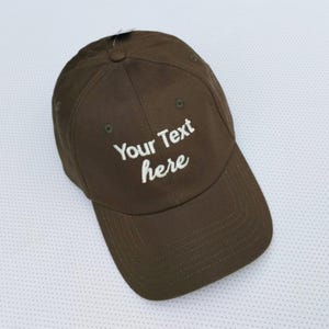 May include: A brown baseball cap with a curved peak. The cap has the text "Your Text here" embroidered in white on the front. It features a button on top and an adjustable strap.