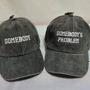 Puede incluir: Dos gorras de béisbol gris carbón con un acabado desgastado y lavado. La gorra de la izquierda presenta el texto bordado en blanco "SOMEBODY" y la de la derecha el texto bordado en blanco "SOMEBODY'S PROBLEM" centrado en los paneles frontales.
