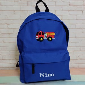 May include: A blue backpack with a black handle and zip. It has an embroidered red fire engine with yellow details. The name "Nino" is embroidered in white on the front.
