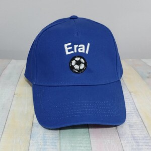 May include: A royal blue baseball cap featuring the word "Eral" in white above a black and white football design. The cap has a curved peak and a structured crown, suitable for sports or casual wear.