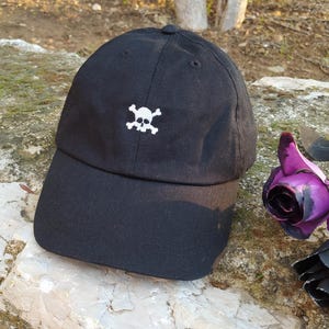 May include: Black baseball cap featuring a white skull and crossbones emblem. The cap has a curved peak and a button on the crown. A purple rose and a black rose are next to the cap.