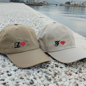 May include: Two baseball caps, one tan and one off-white, are displayed. The tan cap has a black "L" with a red heart, while the off-white cap has a black "F" with a red heart. Both caps have a curved brim.