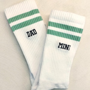 Peut inclure: Paire de chaussettes blanches avec des rayures vertes et les mots "DAD" et "MINI" brodés en noir. Les chaussettes sont côtelées et semblent être faites d'un matériau doux et confortable. Idéales pour un père et son enfant.