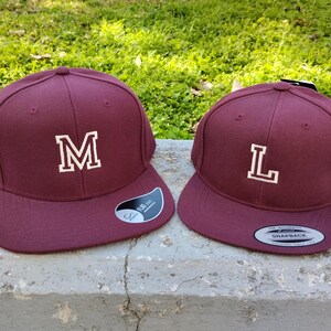 Puede incluir: Dos gorras de béisbol de color burdeos con letras bordadas en blanco. La gorra de la izquierda tiene la letra "M" y la gorra de la derecha tiene la letra "L".