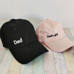 Könnte beinhalten: Zwei Baseballkappen, eine schwarze und eine rosa, werden gezeigt. Auf der schwarzen Kappe ist das Wort "Dad" in Weiß gestickt. Auf der rosa Kappe steht "Dad's girl" in Schwarz gestickt. Beide Kappen haben einen gebogenen Schirm.