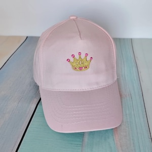 Op de afbeelding: Lichtroze baseballpet met een gebogen klep. De pet heeft een gouden geborduurde kroon met roze en rode details op de voorkant. De pet is gemaakt van een zachte stof en heeft een afgeronde bovenkant.