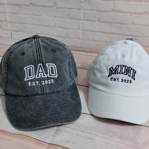 May include: Two baseball caps, one dark gray and one white. The dark gray cap has "DAD EST. 2023" embroidered in white. The white cap has "MINI EST. 2023" embroidered in black. Both caps are casual and likely for a parent and child.