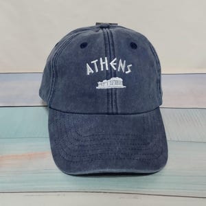 May include: A navy blue baseball cap with a distressed, washed-out finish. The front features white embroidered text that reads ATHENS, positioned directly above a white embroidered graphic of the Parthenon temple structure.