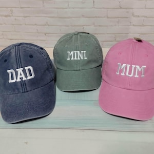 Puede incluir: Tres gorras de béisbol. Una gorra azul marino descolorida con "DAD" bordado en blanco, una gorra verde con "MINI" y una gorra rosa con "MUM". Todas las gorras tienen una visera curva y una correa ajustable.