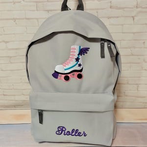 Könnte beinhalten: Ein hellgrauer Rucksack mit einer Vordertasche und einem oberen Griff. Der Rucksack hat ein gesticktes Rollschuh-Design mit rosa, weißen und blauen Akzenten und das Wort "Roller" in lila Schrift.