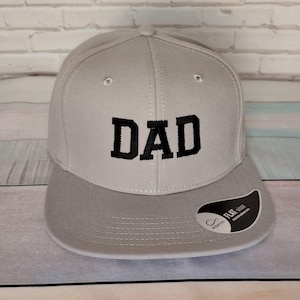 Könnte beinhalten: Eine graue und schwarze Baseballkappe mit dem Wort "DAD" in Schwarz auf der Vorderseite gestickt. Die Kappe hat einen flachen Schirm und eine graue Unterseite.