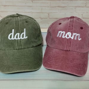 Puede incluir: Dos gorras de béisbol, una verde oliva y otra burdeos. La gorra verde oliva tiene la palabra "dad" bordada en blanco. La gorra burdeos tiene la palabra "mom" bordada en blanco. Ambas gorras tienen una visera curva.