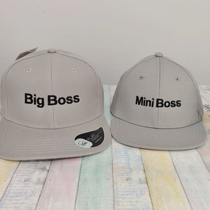 Könnte beinhalten: Zwei graue Baseballkappen mit schwarzer Stickerei. Die größere Kappe trägt die Aufschrift "Big Boss" und die kleinere die Aufschrift "Mini Boss".