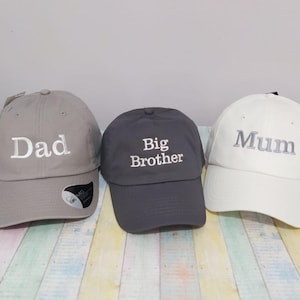 May include: Three baseball caps in grey, dark grey, and white. The grey cap has "Dad" embroidered in white. The dark grey cap has "Big Brother" embroidered in white. The white cap has "Mum" embroidered in grey. All caps have a curved peak.