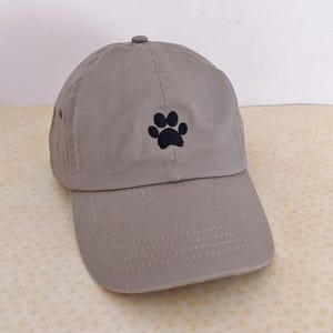 May include: A gray baseball cap with a curved brim. The cap features a black paw print embroidered on the front. The cap has a rounded top and an adjustable strap at the back.