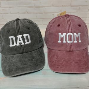 Può includere: Due cappellini da baseball, uno grigio scuro e uno bordeaux. Il cappellino grigio scuro ha la parola "DAD" ricamata in bianco. Il cappellino bordeaux ha la parola "MOM" ricamata in bianco. Entrambi i cappellini hanno un aspetto vintage.