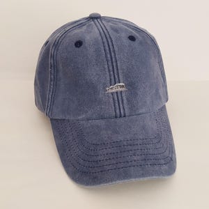 Puede incluir: Una gorra de béisbol azul marino con un acabado de tela desgastada y lavada. El panel frontal presenta un pequeño gráfico bordado en blanco centrado del Partenón. La gorra incluye una visera curva con costuras decorativas.