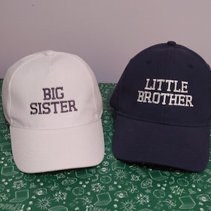 May include: Two baseball caps, one white and one navy blue. The white cap has "BIG SISTER" embroidered in gray, while the navy cap says "LITTLE BROTHER" in white. Both caps have curved visors and adjustable straps.