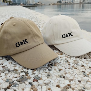 Peut inclure: Deux casquettes de baseball, une beige et une blanc cassé, sont présentées côte à côte. Chaque casquette a une visière incurvée et les lettres brodées "G&K" en noir. Elles reposent sur une surface de petits cailloux clairs.