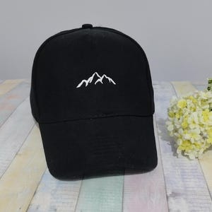 May include: Black baseball cap with a white mountain range embroidered on the front. The cap has a curved brim and a button on top. The hat is sitting on a light-colored wooden surface.