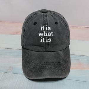 Puede incluir: Una gorra de béisbol gris carbón con un acabado desgastado y lavado. La parte frontal de la gorra presenta el texto bordado en blanco "it is what it is" en una fuente sencilla sin remates. La gorra tiene una visera curva y costuras.