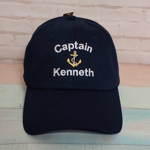 Könnte beinhalten: Marineblaue Baseballkappe mit dem gestickten Text "Captain Kenneth" über einem goldenen Ankeremblem. Die Kappe hat einen gebogenen Schirm und eine strukturierte Krone.