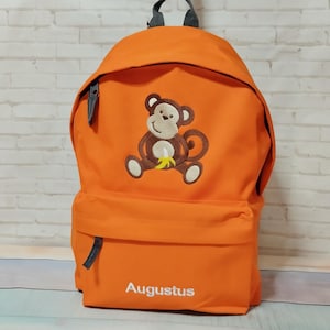 May include: Orange backpack with a monkey design holding a banana. The backpack has a front pocket, a top handle, and the name "Augustus" embroidered in white. The monkey is brown and tan with white accents.
