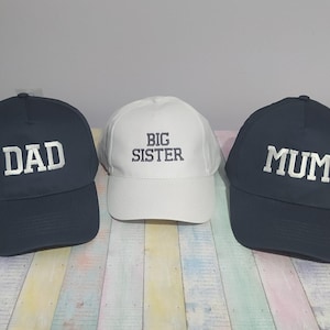 Peut inclure: Trois casquettes de baseball. Deux casquettes bleu marine avec "DAD" et "MUM" brodés en blanc. Une casquette blanche au centre avec "BIG SISTER" brodé en noir. Les casquettes sont posées sur une surface rayée.