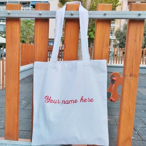 Puede incluir: Bolso tote blanco con asas largas, colgado de una valla de madera. La bolsa presenta el texto bordado "Your name here" en cursiva roja. El fondo muestra un parque con edificios y árboles.