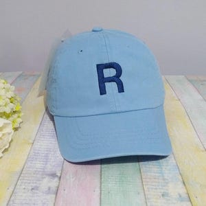 Könnte beinhalten: Eine hellblaue Baseballkappe mit einem dunkelblau gestickten Buchstaben R auf der Vorderseite. Die Kappe hat eine strukturierte Krone, einen gebogenen Schirm mit sichtbaren Nähten und einen Knopf oben.