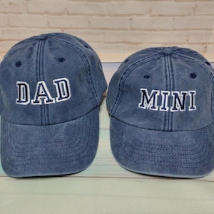 May include: Two blue denim baseball caps. One cap has the word "DAD" embroidered in white, and the other has "MINI" embroidered in white. Both caps have a curved brim and adjustable strap.