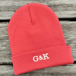 Op de afbeelding: Een fel koraalkleurige gebreide muts met een omgeslagen boord. Op de voorkant van de boord staat de geborduurde witte tekst G&K in een schreeflettertype. De muts is gemaakt van geribbelde gebreide stof met een ronde bovenkant en een zichtbare verticale naad.