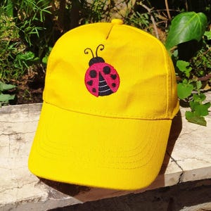 May include: Bright yellow baseball cap featuring an embroidered ladybug design. The ladybug is red with black spots and a black head. The cap has a curved brim and a button on top. A fun accessory for outdoor activities.
