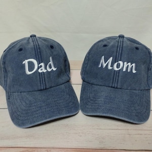 Könnte beinhalten: Zwei Baseballkappen aus blauem Denim mit verwaschener Optik. Die linke Kappe zeigt den weiß gestickten Schriftzug Dad, die rechte Kappe den weiß gestickten Schriftzug Mom. Beide Kappen haben einen gebogenen Schirm und eine Sechs-Panel-Konstruktion.