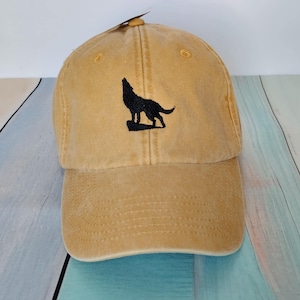 May include: Mustard-colored baseball cap with a curved brim. The cap features a black embroidered silhouette of a howling wolf on the front. The cap has a classic six-panel design with a top button and adjustable strap.