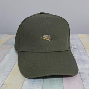 Op de afbeelding: Olijfgroene baseballpet met een klein geborduurd beverontwerp op de voorkant. De pet heeft een gebogen klep en een knoop aan de bovenkant. De achtergrond is een lichtgekleurd houten oppervlak.