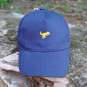 Könnte beinhalten: Marineblaue Baseballkappe mit einem gelben, gestickten Dinosaurier-Logo auf der Vorderseite. Die Kappe hat einen gebogenen Schirm und einen Metallverschluss auf der Rückseite. Hergestellt aus strapazierfähigem Stoff.