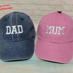 Puede incluir: Un conjunto de dos gorras de béisbol con el texto Set of two baseball caps. Una gorra es azul oscuro con el texto bordado en blanco DAD, y la otra es rosa con el texto bordado en blanco MUM. Ambas gorras tienen una visera curva y costuras.