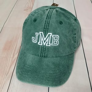 Könnte beinhalten: Eine grüne Baseballkappe mit den gestickten Buchstaben "JMB" in Weiß. Die Kappe hat einen gebogenen Schirm und einen Used-Look im Vintage-Stil. Der Hut ist aus einem weichen, strukturierten Stoff gefertigt.
