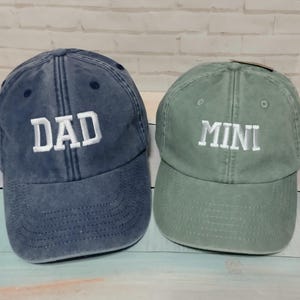 Può includere: Due cappellini da baseball, uno blu navy e uno verde salvia. Il cappellino blu navy ha la scritta "DAD" ricamata in bianco, e il cappellino verde ha "MINI" ricamato in bianco. Entrambi i cappellini hanno una visiera curva e un cinturino regolabile.