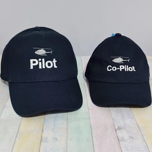 May include: Two navy blue baseball caps. The larger cap has the word "Pilot" embroidered in white, with a helicopter above the word. The smaller cap has the words "Co-Pilot" embroidered in white, with a helicopter above the words.