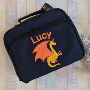May include: A navy blue lunch bag with a black trim and handle. The bag features the name "Lucy" in orange, above an orange and yellow dragon design. The bag is rectangular and has a zip closure.