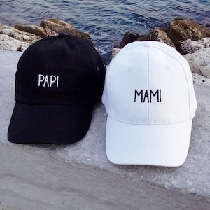 Puede incluir: Se muestran dos gorras de béisbol, una negra y otra blanca. La gorra negra tiene la palabra "PAPI" bordada en blanco, mientras que la blanca tiene "MAMI" bordada en negro. Ambas gorras están sobre una superficie de piedra.