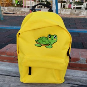 Könnte beinhalten: Ein leuchtend gelber Rucksack mit einem schwarzen Griff und Trägern. Der Rucksack hat eine grüne Schildkröten-Grafik auf der Vordertasche. Er hat eine Vordertasche und einen schwarzen Reißverschluss.