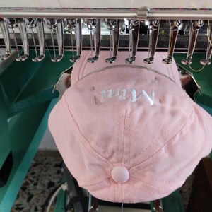 Puede incluir: Gorra de béisbol rosa con la palabra "Mim" bordada en hilo blanco. La gorra está siendo bordada por una máquina con múltiples agujas e hilos. La máquina está sobre un fondo verde y plateado.